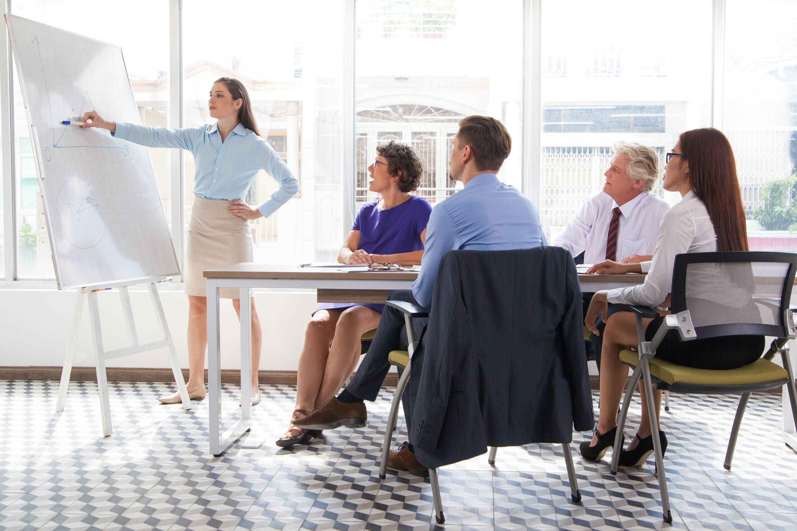 Confident female business coach pointing at whiteboard and explaining graph at meeting, group of four people listening to her. They sitting in conference room. Business training or education concept